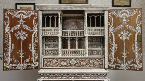 A dark wood cabinet, inlaid with ivory in a design of flowers, leaves and scrolls. It is on a stand, with the doors open to show an interior of drawers and columns, also inlaid with ivory.